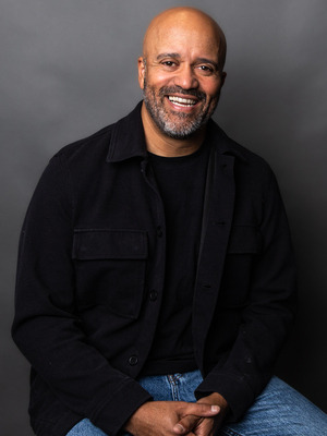 headshot of carlos whittaker with wide smile, black shirt and jacket, and grey background.